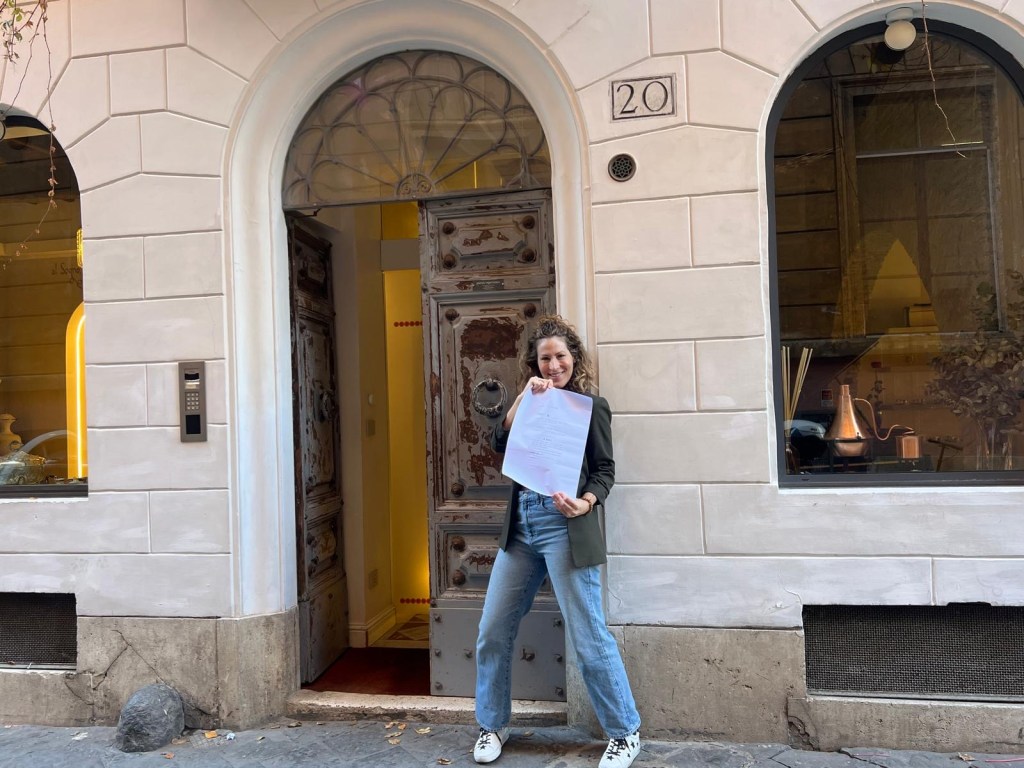 Image of a woman smiling and holding a long sheet of white paper while leaning on a white brick wall near an open door.